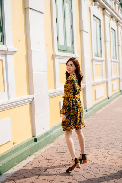 Χαριτωμένο σγουρό μακρύ κορίτσι μαλλιά σε κίτρινο φόρεμα με τα πόδια κατά μήκος του παστέλ κτιρίου στο φως του ήλιου. - Φωτογραφία, εικόνα