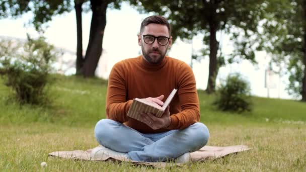 Full shot of young bearded Caucasian man wearing jeans, sweater and glasses, sitting with legs crossed on blanket over green lawn, reading book, then seeing invisible friend, greeting and talking - Footage, Video