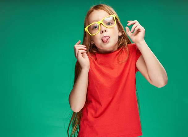 χαρούμενα κόκκινα μαλλιά κορίτσι σε ένα κόκκινο μπλουζάκι κίτρινα γυαλιά συναισθήματα παιδική ηλικία - Φωτογραφία, εικόνα