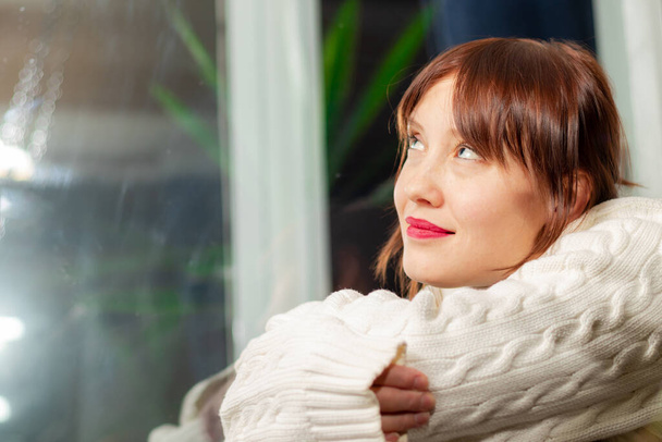 młoda ładna dziewczyna w białym swetrze siedząca na parapecie w domu przy oknie marząc - Zdjęcie, obraz