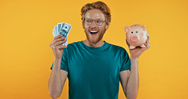 Kumbarayı elinde tutan mutlu kızıl saçlı adam ve para sarıda izole edilmiş. - Fotoğraf, Görsel