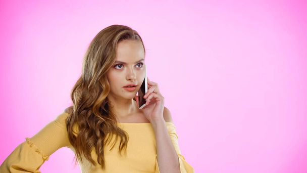 髪の毛が波打つ若い女性がピンクに隔離されたスマートフォンで話している  - 写真・画像