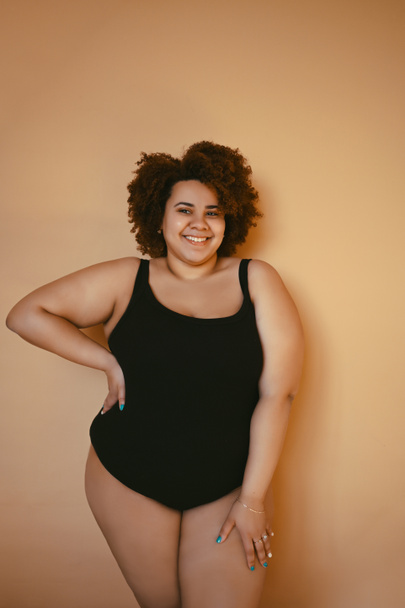 Beautiful curvy oversize African black woman afro hair posing in black bodysuit on beige brown background isolated, body imperfection, body acceptance, body positive and diversity concept. Copyspace. - Photo, Image