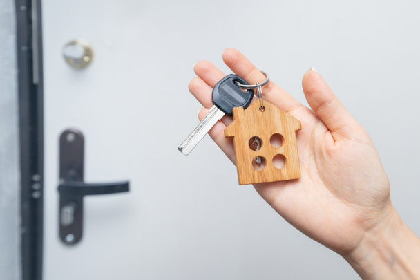 Die Frau hält den Wohnungsschlüssel mit einem Schlüsselanhänger aus Holz im Hintergrund der Eingangstür zur Wohnung. Das Konzept des Umzugs in ein neues Zuhause - Foto, Bild