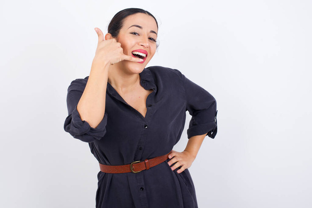 Arab woman wearing dress against white studio background imitates telephone conversation, makes phone call gesture with hands, has confident expression. Call me! - Photo, Image