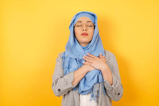 Faithful Young beautiful muslim woman wearing hijab  closes eyes and keeps hands on chest near heart, shows her kindness or favour, expresses sincere emotions, being kind hearted and honest. - Foto, Imagem