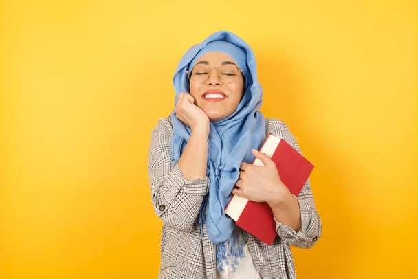 Portret młodej muzułmanki noszącej hidżab trzymającej książkę przytłoczoną emocjami, wyrażającej emocje i szczęście zamkniętymi oczami i dłonią przy twarzy, uśmiechającej się szeroko na jasnym tle. - Zdjęcie, obraz