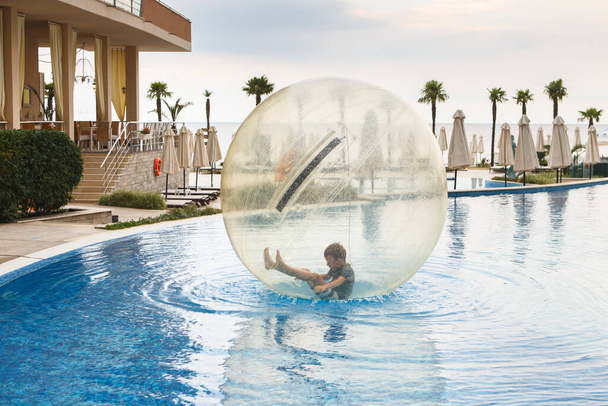 Kind veel plezier in grote plastic ballon op het water van het zwembad op het zomerresort. Kleine jongen in grote opblaasbare transparante bal lopen en plezier hebben.  - Foto, afbeelding