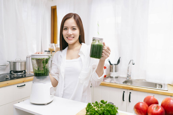 Portré gyönyörű ázsiai nő, hogy smoothie friss zöldségekkel turmixgép és nézi a kamera a konyhában otthon. Egészséges táplálkozás, diéta és életmód koncepció. Vegetáriánus kaja. Szépség Táplálkozás koncepció. - Fotó, kép