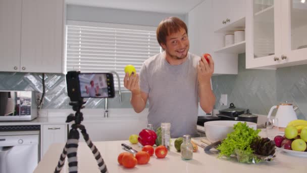 Food blogger cooking meal and recording video for blog. Young cook looking at camera filming himself on smartphone for culinary blog while preparing healthy meal with vegetables in kitchen. - Footage, Video
