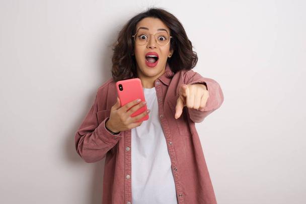 Nuori kaunis nainen älypuhelimella osoittaa studiossa. Arabi nainen yllään lasit ja vaaleanpunainen takki tilalla matkapuhelin vastaan valkoinen seinä - Valokuva, kuva