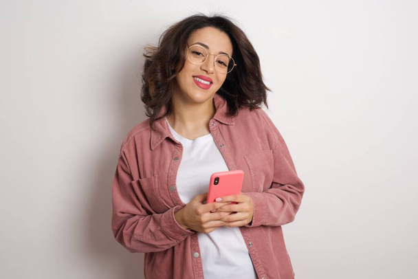 Νεαρή όμορφη γυναίκα που κρατά smartphone στο στούντιο. Αραβίδα γυναίκα φορώντας γυαλιά και ροζ μπουφάν κρατώντας το κινητό τηλέφωνο στον λευκό τοίχο - Φωτογραφία, εικόνα