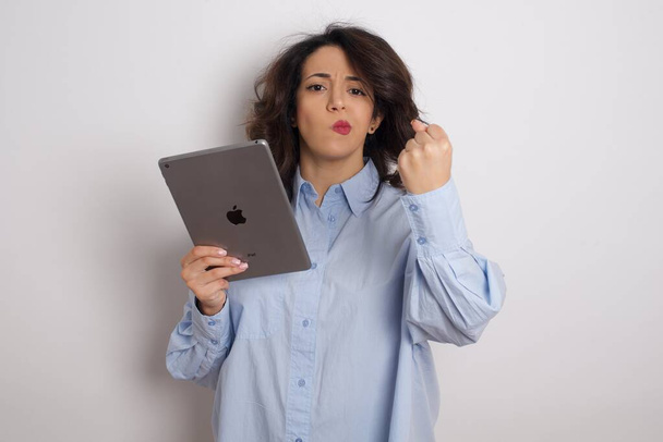 joven hermosa mujer de negocios vistiendo camisa azul sobre la pared blanca con la tableta en el estudio mostrando puño  - Foto, Imagen