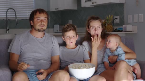 Ευτυχισμένη νεαρή ευρωπαϊκή οικογένεια με δύο παιδιά συγκεντρώνονται στον καναπέ για να παρακολουθήσουν τηλεόραση στο σπίτι χαμογελώντας. Συναισθήματα έκπληξης. Συγκέντρωση και ψυχαγωγία. - Πλάνα, βίντεο