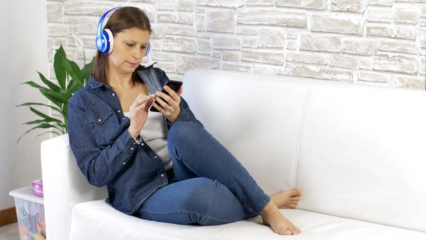 Mulher bonita vestida de jeans ouvindo música com fones de ouvido no sofá branco em casa - Foto, Imagem
