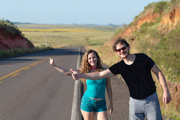 Ένα νεαρό ζευγάρι Βραζιλιάνων κάνει ωτοστόπ στην άκρη ενός αυτοκινητόδρομου. Ταξίδια και τουρισμός. Τρόπος ζωής. Ταξίδι με σακίδιο. αγροτικό τοπίο. - Φωτογραφία, εικόνα