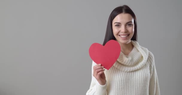 Gyönyörű fiatal nő piros papír szív fúj csók szürke háttér - Felvétel, videó