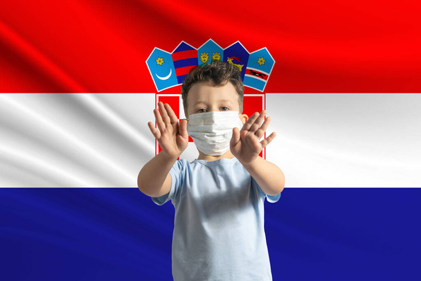 Μικρό λευκό αγόρι με προστατευτική μάσκα στο φόντο της σημαίας της Κροατίας. Κάνει στοπ με τα χέρια του, μένει σπίτι Κροατία. - Φωτογραφία, εικόνα