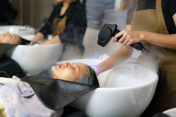 Peluquería está utilizando Nano Mist Hair Treatment Streamer Machine, equipo moderno para joven feliz asiático hermosa mujer caucásica cliente en silla de champú en el salón de pelo. Concepto de belleza. - Foto, imagen