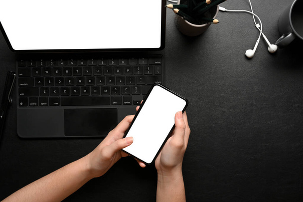 Overhead-Aufnahme weiblicher Hände mit Smartphone auf dunklem kreativen Arbeitsplatz mit Laptop und Zubehör, Clipping-Pfad - Foto, Bild