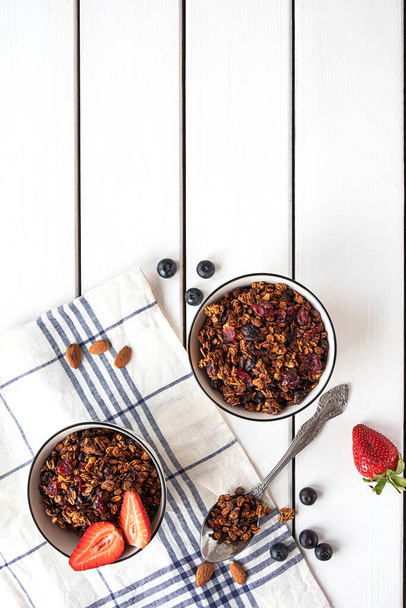 Home-made baked granola in two bowls, with berries, almonds, textile napkin and a spoon. White rustic background, copy space.  - Photo, Image