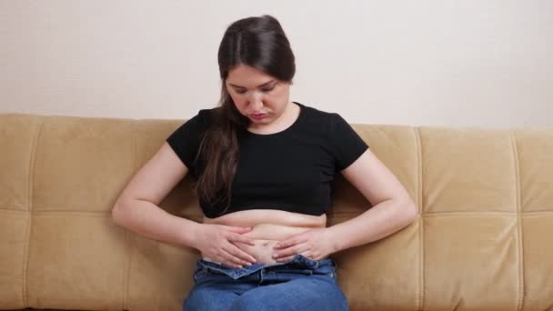 Mujer joven sintiendo pliegues de grasa en el estómago mientras se sienta en el sofá - Imágenes, Vídeo