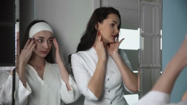 Молодая женщина-косметолог консультирует клиентку, которая смотрит в зеркало в клинике красоты. - Кадры, видео