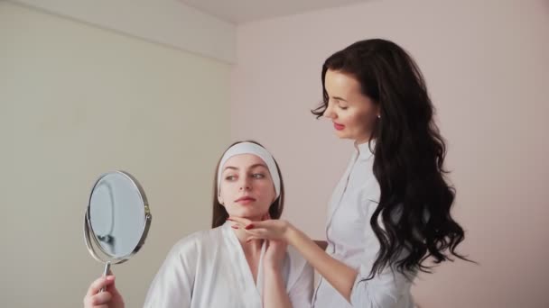Portret van jonge vrouw patiënt die kijkt naar spiegel na gezicht huidreiniging procedure en praten over resultaat met schoonheidsspecialiste in schoonheidskliniek. Behandeling en genezing door cosmetoloog in de kosmetologie. - Video