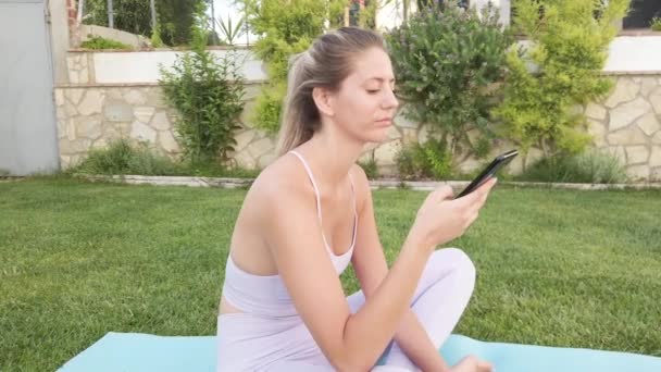 Calma femmina in abbigliamento sportivo seduto a piedi nudi sul tappeto utilizzando il telefono cellulare durante la pratica dello yoga - Filmati, video