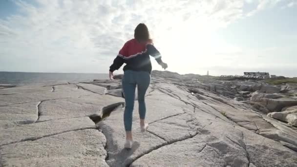 Обратный вид анонимной туристки, бегущей по бурному каменистому побережью против серого облачного неба около моря в бухте Пеггис, Канада - Кадры, видео