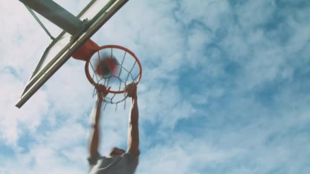 From below ethnic sportsman performing slam dunk shot and hanging on hoop against cloudy sky during basketball training on street - Footage, Video