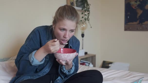 Pensive barefoot female in true wireless earphones and casual clothes with bowl of tasty food focusing on screen with interest while sitting with legs crossed on soft bed and using netbook in cozy apartment - Footage, Video