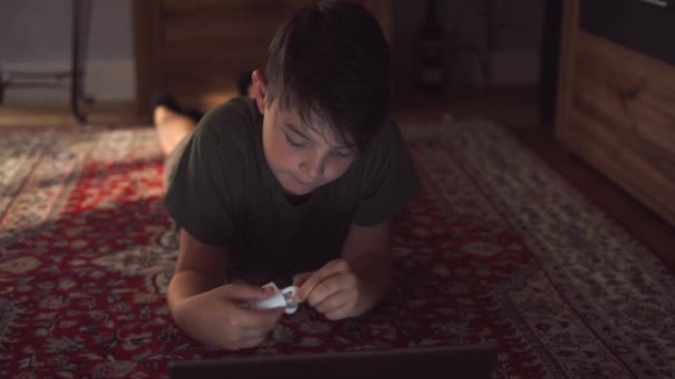 Pensive malý chlapec v neformální oblečení ležící na podlaze a dávat na sluchátka a prohlížení notebook během víkendu doma - Záběry, video
