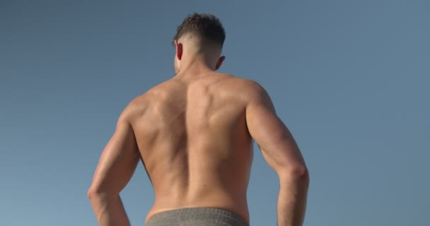 Takaisin näkymä lihaksikas mies kehonrakentaja alaston vartalo seisoo meren rannalla ja tekee harjoituksia vastus bändi harjoituksen aikana kesällä - Materiaali, video