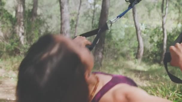 Zadní pohled na anonymní sportovkyně ve sportovním oblečení tahání nahoru s TRX popruhy přivázané ke stromu při provozování sportu a stojící na zeleném trávníku za slunečného letního dne v lese - Záběry, video