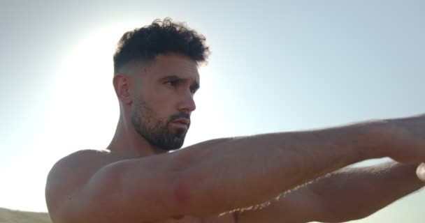 Muscular male bodybuilder with naked torso standing on seashore and doing exercises with resistance band during workout in summer - Footage, Video