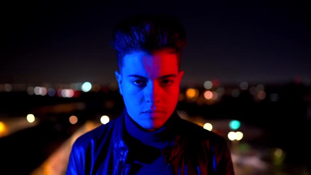 Nykyaikainen nuori kaveri peittää kasvot käsin seisoessaan kirkkaan punaisen ja sinisen valon alla kaupungin kadun hämärtyneellä taustalla yöllä - Materiaali, video