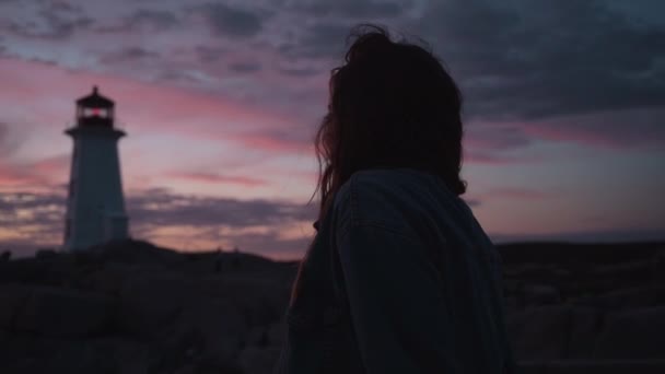 Mladá turistka v džínové bundě se směje, zatímco stojí na rozmazaném pozadí oblačného západu slunce a Peggys Cove Lighthouse ve večerních hodinách v Kanadě - Záběry, video