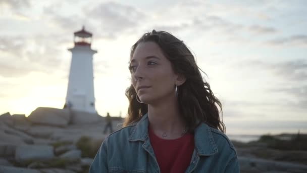 Young female tourist in denim jacket laughing while standing on blurred background of cloudy sundown sky and Peggys Cove Lighthouse in evening in Canada - Footage, Video