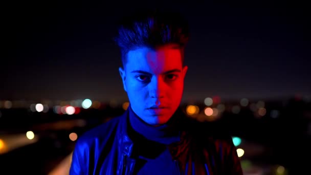 Contemporary young guy covering half of face with hand and looking at camera while standing under bright red and blue light on blurred background of city street at night - Footage, Video