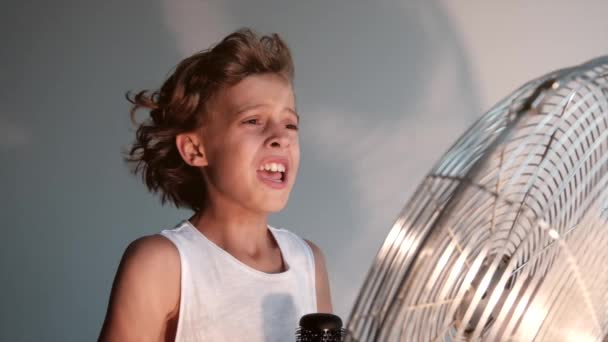 Bambino con espressione emotiva che canta con una spazzola per capelli come un microfono davanti a un tavolo con un ventilatore che rimuove i capelli all'interno di una stanza - Filmati, video