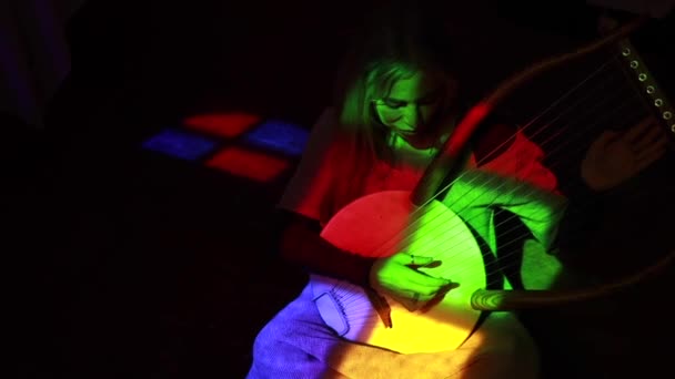 Z góry blondynka gra tradycyjną muzykę na lirze siedząc w pokoju z wielobarwnym oświetleniem - Materiał filmowy, wideo