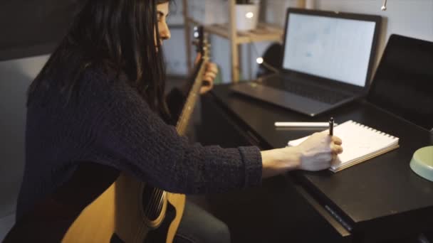 Vista lateral da fêmea tocando guitarra acústica ao compor música perto da mesa com laptop no quarto com parede de tijolo durante o trabalho remoto - Filmagem, Vídeo