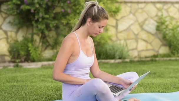 Mujer tranquila en ropa deportiva sentada descalza en la esterilla y eligiendo un tutorial en línea para practicar yoga - Imágenes, Vídeo