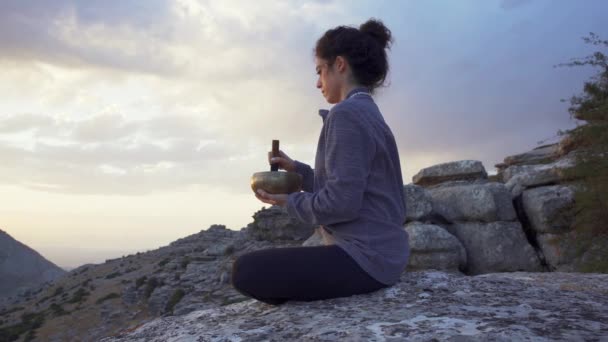 Low angle side view of tranquil female sitting in Padmasana position with singing bowl and meditating while relaxing in mountains at sunset time - Footage, Video