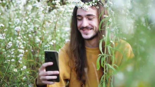 Romantikus fiatal, hosszú hajú férfi alkalmi ruhában ül virágzó vadvirágok között, és szelfit készít a mobillal, miközben nyaranta vidéken pihen. - Felvétel, videó