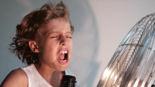 Gyermek érzelmi kifejezés énekel egy hajkefével, mint egy mikrofon előtt egy asztal egy ventilátor, amely eltávolítja a hajat a szobában - Felvétel, videó