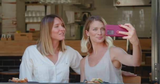 Експресивні подружки посміхаються, приймаючи селфі зі смартфоном в кафе, сидячи біля відкритого вікна
 - Кадри, відео