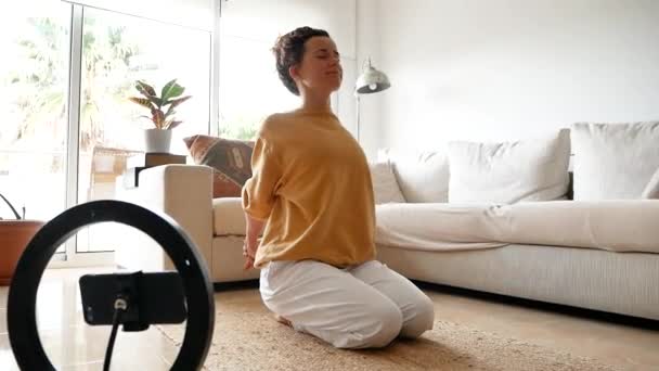 Mujer tranquila en ropa deportiva sentada con los ojos cerrados en el suelo con teléfono móvil haciendo tutorial en pose vajrasana mientras practica yoga en casa - Imágenes, Vídeo