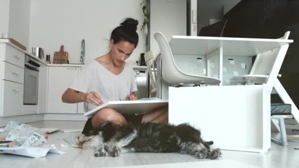 Mujer joven concentrada sentada en el suelo cerca del perro y leyendo instrucciones de papel mientras instala muebles nuevos en un apartamento moderno - Imágenes, Vídeo